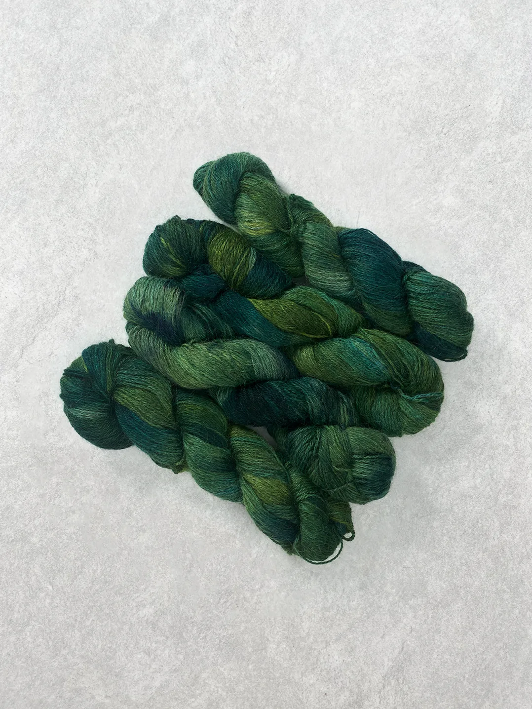 Twisted skeins of hand-dyed wool yarn in variegated shades of deep emerald and forest green on a textured background